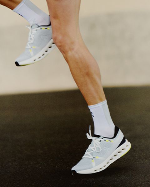 Eine Nahaufnahme einer Person beim Laufen, die ihre Unterschenkel in weißen Sportschuhen mit schwarzen und gelben Akzenten und weißen Socken vor einem unscharfen Hintergrund zeigt.