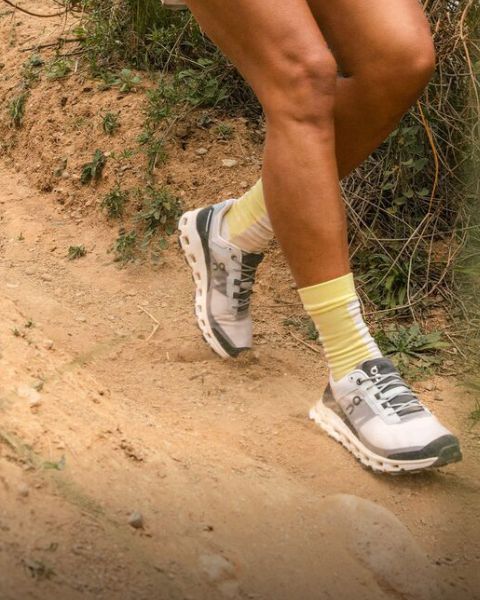 Eine Person, die gelb gestreifte Socken und weiße Laufschuhe trägt, läuft auf einem Feldweg, der von Gras und kleinen Pflanzen umgeben ist. Nur die Unterschenkel und Füße sind sichtbar.