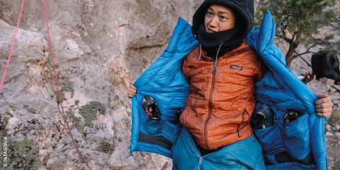 Eine Person im Freien trägt eine orangefarbene Daunenjacke, die geöffnet ist und eine blaue Jacke darunter zum Vorschein bringt. Sie richtet ihre Kleidung, umgeben von felsigem Gelände und Kletterausrüstung. In weißem Text steht „DAUNENJACKEN PFLEGEN“ mit einem Logo auf der rechten Seite.