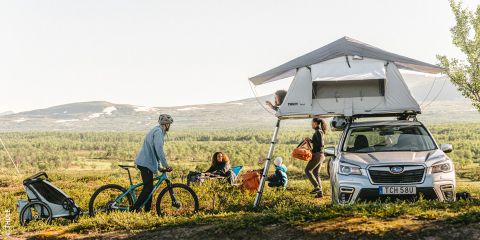 Eine Gruppe von Menschen campt in einer malerischen Berggegend. In der Nähe parkt ein Auto mit Dachzelt, daneben ein Fahrrad mit Anhänger. Die Menschen genießen die Natur unter einem klaren Himmel. Der deutsche Text lautet „Nachhaltige Wandertipps“.