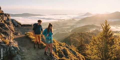 Zwei Menschen wandern bei Sonnenaufgang auf einem Bergpfad, umgeben von zerklüftetem Gelände und nebelverhangenen Tälern. Das warme Sonnenlicht taucht die Landschaft in einen goldenen Schimmer. Der Text lautet „Wanderschuh Guide“ mit einem Hinweis auf Dachstein.