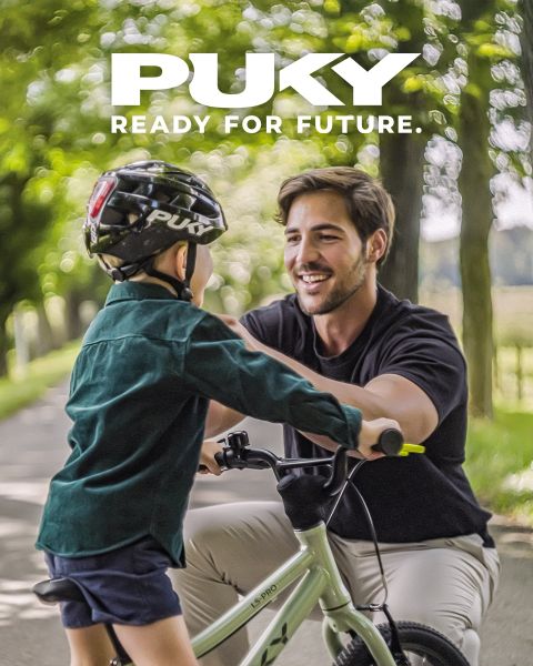 Ein junges Mädchen mit blauem Helm und gestreiftem Shirt fährt an einem sonnigen Tag auf einem hellblauen Fahrrad. Lächelnd fährt sie an bunten Graffitis in einer urbanen Umgebung vorbei. Das PUKY-Logo und der Slogan „Ready for life“ sind oben links zu sehen.