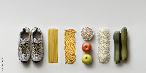 Ein Apfel, eine Zucchini und ungekochte Nudeln liegen auf einer Fläche, daneben Haferflocken. Im Hintergrund steht der deutsche Text: „ERNÄHRUNG IM LAUFSPORT“, was auf die Ernährung im Laufsport hindeutet.