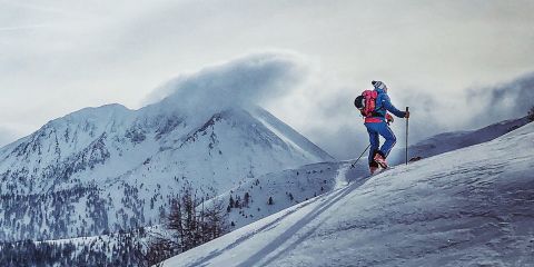 Eine Person in Winterkleidung fährt einen verschneiten Berghang hinauf, im Hintergrund ein bewölkter Himmel und entfernte Berggipfel. Der Text auf Deutsch lautet „PACKLISTE FÜR DIE SKITOUR“.