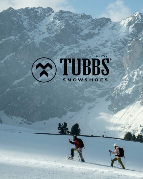 Zwei Personen, die mit Skistöcken einen verschneiten Hang hinaufgehen, vor dem Hintergrund hoher, schroffer, schneebedeckter Berge. Das Tubbs Snowshoes-Logo erscheint über dem Bild.
