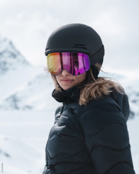 Eine Person, die einen schwarzen Skihelm, eine reflektierende Skibrille und eine dunkle Pufferjacke trägt, steht in einer verschneiten Berglandschaft, mit schneebedeckten Gipfeln im Hintergrund.