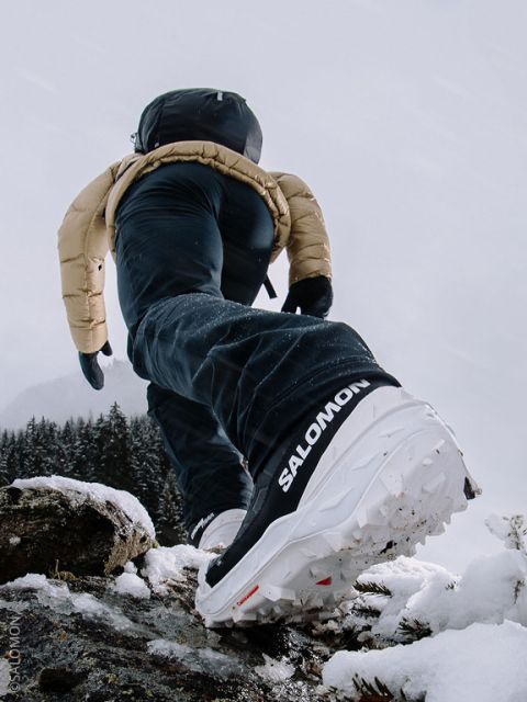 Eine Person in Winterkleidung und Salomon-Schuhen schreitet auf Schnee bergauf, wobei der Schwerpunkt auf der Schuhsohle vor einem verschneiten Berghintergrund liegt.