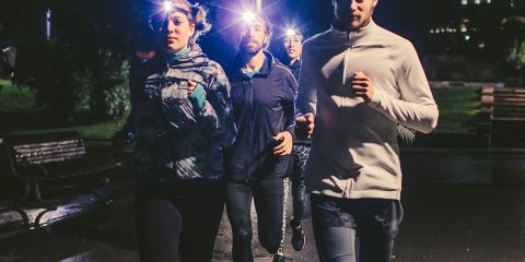 Vier Personen joggen nachts mit Stirnlampen auf einem schwach beleuchteten Weg. Im Hintergrund sind beleuchtete Gebäude zu sehen. Der Text lautet „Sicher & Sichtbar Laufen im Dunkeln“ und weist auf die Sichtbarkeit beim Laufen im Dunkeln hin.