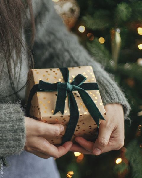 Eine Person in einem grauen Pullover hält ein in Goldpapier mit Sternenmustern eingewickeltes und mit einem dunkelgrünen Samtband verschnürtes Geschenk vor einem geschmückten Weihnachtsbaum mit Lichtern.