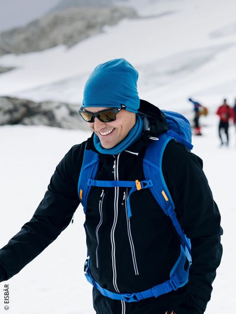 Eine lächelnde Person in Winterkleidung wandert in verschneitem Gelände, trägt eine Sonnenbrille, eine blaue Mütze und einen blauen Rucksack, während im Hintergrund andere Personen zu sehen sind.
