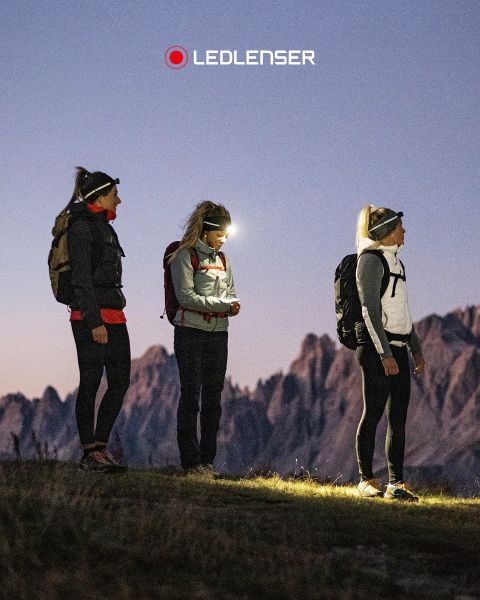 Drei Frauen in Outdoor-Ausrüstung und mit Stirnlampen stehen in der Dämmerung auf einem Grashügel mit Bergen im Hintergrund. Über ihnen das Logo "LEDLENSER".