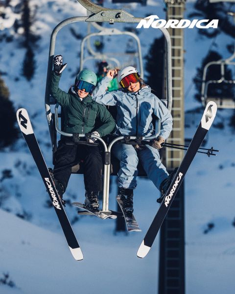 Zwei Skifahrer in Winterausrüstung fahren lächelnd und winkend mit Nordica-Skiern in einem Skilift, im Hintergrund sind verschneite Berge und Bäume zu sehen.