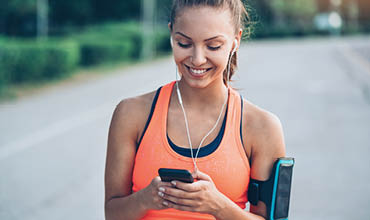 Eine Frau in einem leuchtend orangefarbenen Tanktop joggt im Freien. Sie trägt Kopfhörer und lächelt, während sie auf ihr Smartphone schaut, das sie in einer Hand hält. An ihrem anderen Arm ist eine Handyhalterung befestigt. Hinter ihr sind ein verschwommener Pfad und Grün zu sehen.