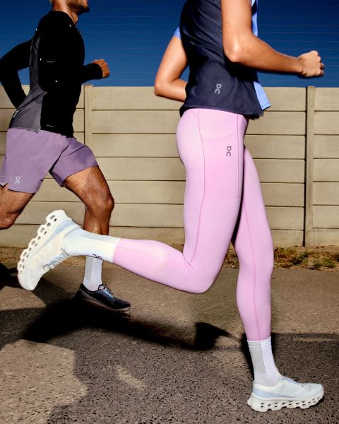 Eine Person läuft im Freien. Sie trägt eine beige-graue Windjacke, hellgraue Shorts und weiße Laufschuhe. Der Hintergrund ist ein klarer Himmel und eine ebene Fläche. Das Logo „On“ ist in der oberen rechten Ecke sichtbar.