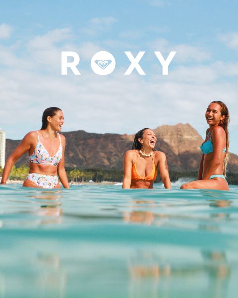 Drei Frauen schwimmen in Badeanzügen unter Wasser in klarem, blauem Wasser. Eine trägt ein Langarmshirt, die anderen Bikinis. Das Bild ist mit dem weißen „Roxy“-Logo überlagert.