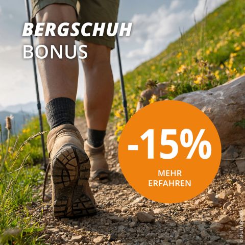 Person mit Rucksack wandert unter leicht bewölktem Himmel einen felsigen Bergpfad hinauf. Der Text lautet „BERGSCHUH BONUS“ und „-15 % MEHR ERFAHREN“ in einem orangefarbenen Kreis. Im Hintergrund sind schneebedeckte Gipfel zu sehen.