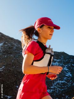 Eine Frau in roter Laufkleidung joggt mit einer Trinkweste unter blauem Himmel auf einer Bergstrecke.