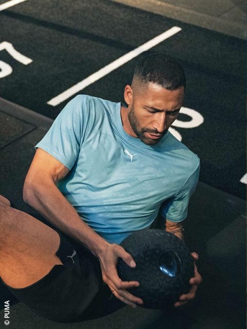 Ein Mann in einem hellblauen Puma-Shirt trainiert auf dem Boden eines Fitnessstudios und hält mit beiden Händen einen schwarzen, strukturierten Medizinball.