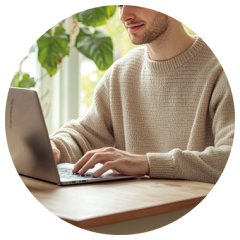 Eine Person in einem beigen Pullover tippt an einem Laptop an einem Holztisch, im Hintergrund sind grüne Pflanzen und Sonnenlicht zu sehen.