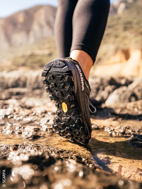 Nahaufnahme einer Person in schwarzen Trailrunning-Schuhen, die auf nassem, felsigem Untergrund im Freien läuft, mit Bergen im unscharfen Hintergrund.