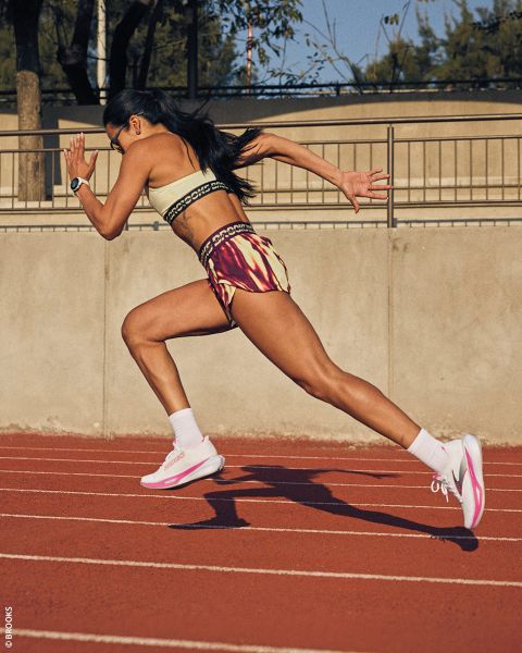 Eine Frau in Sportkleidung sprintet auf einer roten Laufbahn im Freien, die Arme pumpen und der Körper beugt sich mitten im Schritt nach vorne.
