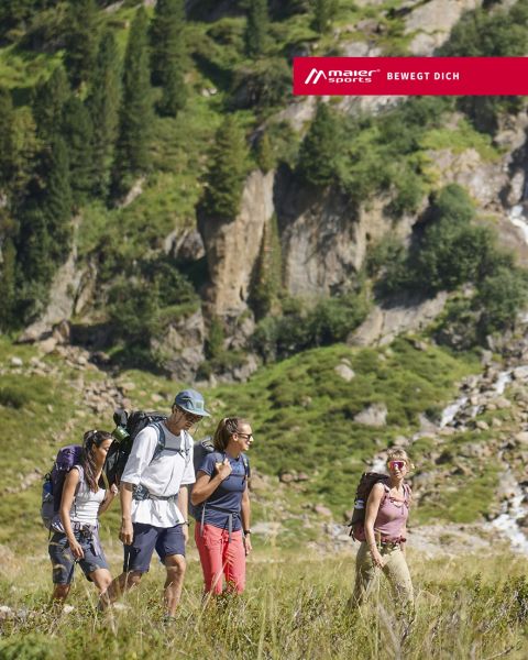 Drei Personen in Winterkleidung wandern unter strahlendem Sonnenschein einen schneebedeckten Berghang hinauf. Sie benutzen Skistöcke und tragen Rucksäcke. Im Hintergrund sind schneebedeckte Gipfel in der Ferne zu sehen.