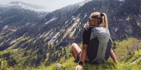 Eine Frau mit einem Rucksack sitzt auf einem grasbewachsenen Hügel und blickt auf eine malerische Berglandschaft mit Wäldern und Felsen.