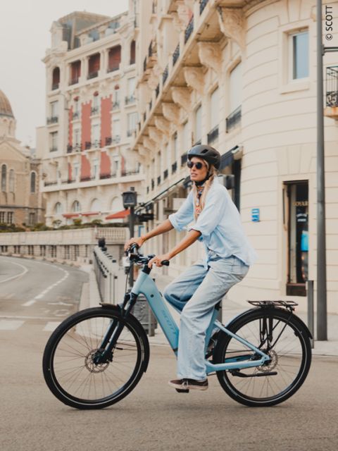 Eine Frau mit Helm und Sonnenbrille fährt auf einem blauen Fahrrad durch eine von eleganten Gebäuden gesäumte Straße.