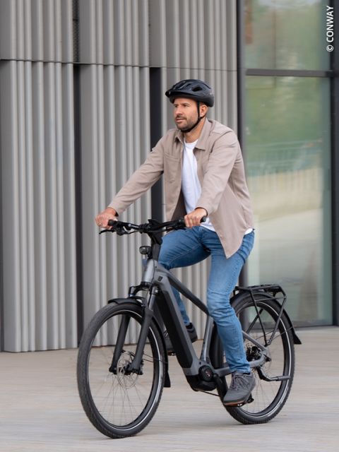 Ein Mann, der einen Helm und Freizeitkleidung trägt, fährt mit einem schwarzen Fahrrad auf einem gepflasterten Platz in der Nähe eines modernen Gebäudes.