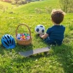 Kleiner Junge sitzt mit einem Fußball im Gras. In der Nähe stehen ein Korb mit bunten Eiern, ein blauer Helm und ein Skateboard. Sonnige grüne Landschaft.