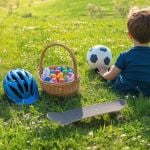 Kleiner Junge sitzt mit einem Fußball im Gras. In der Nähe stehen ein Korb mit bunten Eiern, ein blauer Helm und ein Skateboard. Sonnige grüne Landschaft.