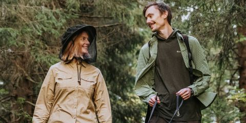 Zwei Menschen, die in einem Wald spazieren gehen und sich anlächeln; der eine trägt eine hellbraune Jacke mit einem Mückennetz, der andere eine grüne Jacke, beide halten Wanderstöcke.