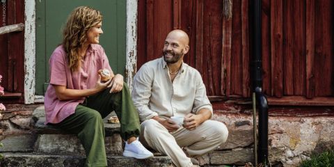 Eine Frau und ein Mann sitzen auf Steinstufen vor einem rustikalen Holzgebäude, lächeln und genießen gemeinsam Kaffee und Gebäck.