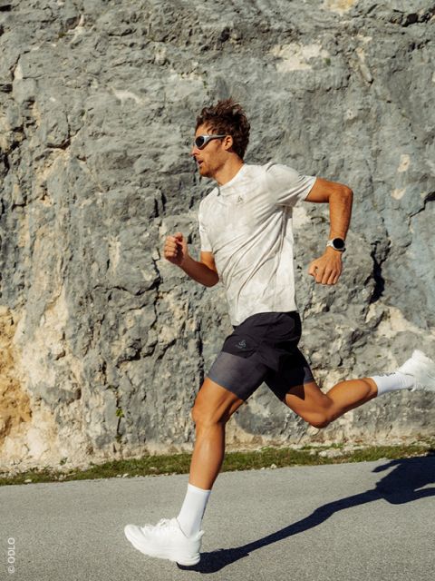 Ein Mann mit Sonnenbrille und sportlicher Kleidung läuft im hellen Sonnenlicht eine Straße entlang, die an einer Felswand vorbeiführt.