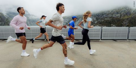 Fünf Personen in Sportkleidung joggen im Freien auf einem gepflasterten Weg, mit Bergen und Wolken im Hintergrund.
