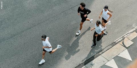 Vier Personen laufen zusammen auf einer gepflasterten Straße, von oben gesehen, in der Nähe eines Bordsteins mit aufgemalten Markierungen.