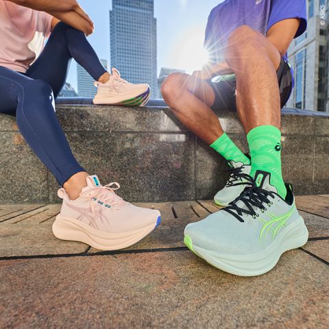 Zwei Personen in Sportkleidung sitzen im Freien auf einer Treppe und zeigen pastellfarbene und neonfarbene ASICS-Laufschuhe, während im Hintergrund die Skyline der Stadt zu sehen ist.
