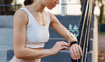 Eine Person joggt auf einer Kopfsteinpflasterstraße, umgeben von hohen Gebäuden. Die Sonne geht unter und wirft einen warmen Schein, während eine blaue Lichtspur hinter ihr herzieht. Der Himmel ist klar und hell.
