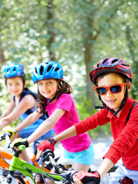Drei Kinder mit Helmen fahren an einem sonnigen Tag im Freien Fahrrad und lächeln in die Kamera. Der Hintergrund ist mit grünen Bäumen gefüllt.
