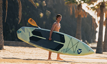 Ein Mann in Badehose läuft mit einem Paddleboard und einem Paddel über einen Sandstrand, mit Palmen und Sonnenlicht im Hintergrund.