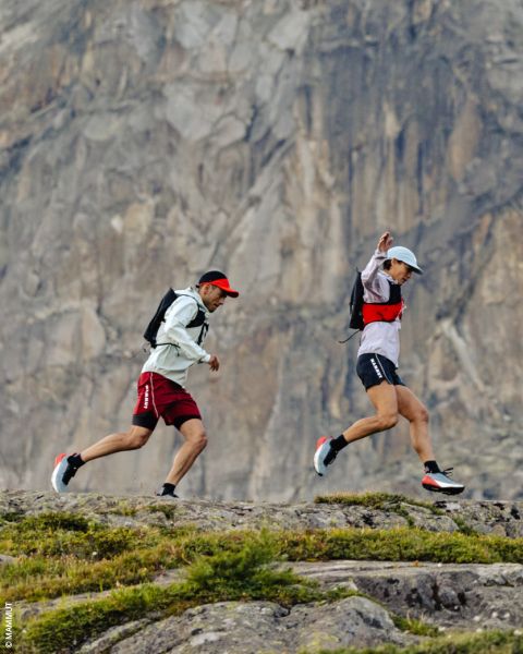 Zwei Personen laufen auf einem felsigen Pfad vor einer steilen Bergklippe, sie tragen Hüte, Rucksäcke und Sportkleidung.