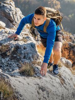 Ein junger Mann in einem blauen Sweatshirt und mit einem Rucksack klettert einen felsigen Berghang hinauf, umgeben von Herbstlaub.