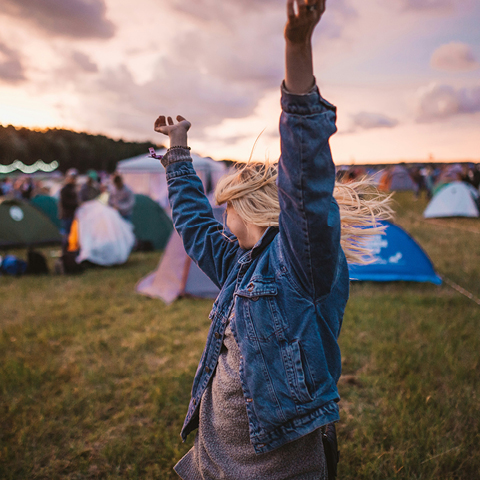 Eine Person in Jeansjacke hebt freudig die Arme auf einem Campingplatz mit Zelten bei Sonnenuntergang.