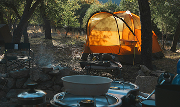 Ein leuchtend orangefarbenes Zelt steht auf einem bewaldeten Campingplatz mit Kochtöpfen und Vorräten auf einem Tisch im Vordergrund.