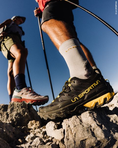 Zwei Wanderer stehen mit Trekkingstöcken auf felsigem Gelände; der Fokus liegt auf dem einen, der schwarze La Sportiva-Schuhe und weiße Socken unter einem klaren blauen Himmel trägt.