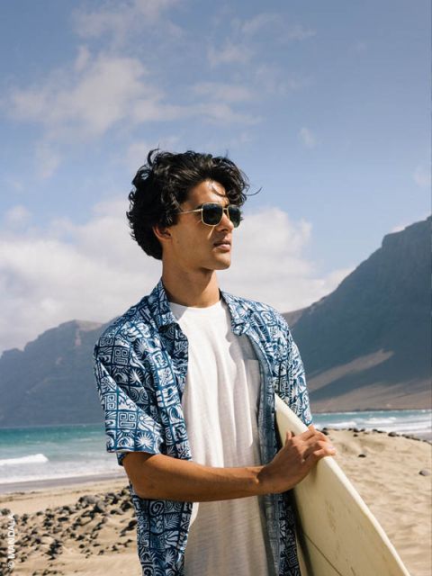 Mann mit Sonnenbrille und blau gemustertem Hemd hält ein Surfbrett in der Hand und steht an einem Sandstrand mit Bergen und Meer im Hintergrund.