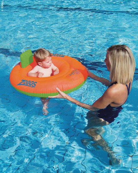 Eine Frau hält ein Baby im Arm, das in einem orange-grünen Schwimmring in einem Schwimmbecken sitzt.