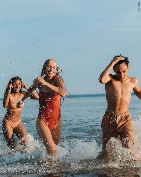 Drei junge Erwachsene in Badekleidung lachen und planschen, während sie an einem sonnigen Tag durch flaches Meerwasser laufen.