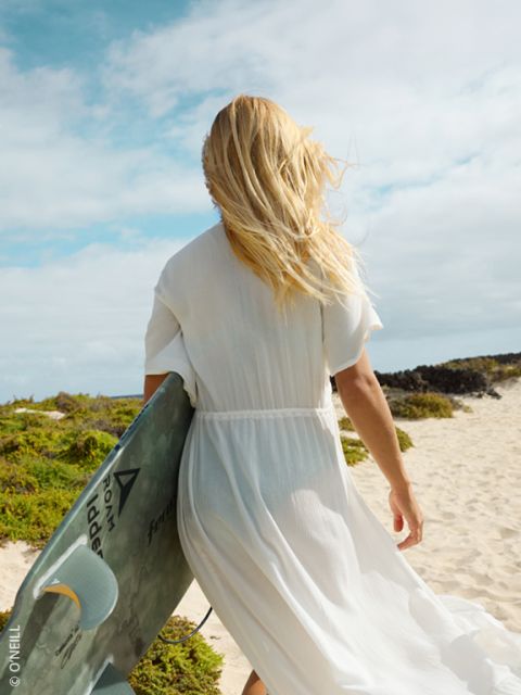 Eine Frau in einem weißen Kleid läuft mit einem Surfbrett in der Hand über einen Sandstrand, ihr blondes Haar weht im Wind.