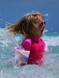 Ein Mädchen mit rosafarbener Sonnenbrille und Badeanzug plantscht im Meer und lächelt aufgeregt, als das Wasser an einem sonnigen Tag um sie herum spritzt.